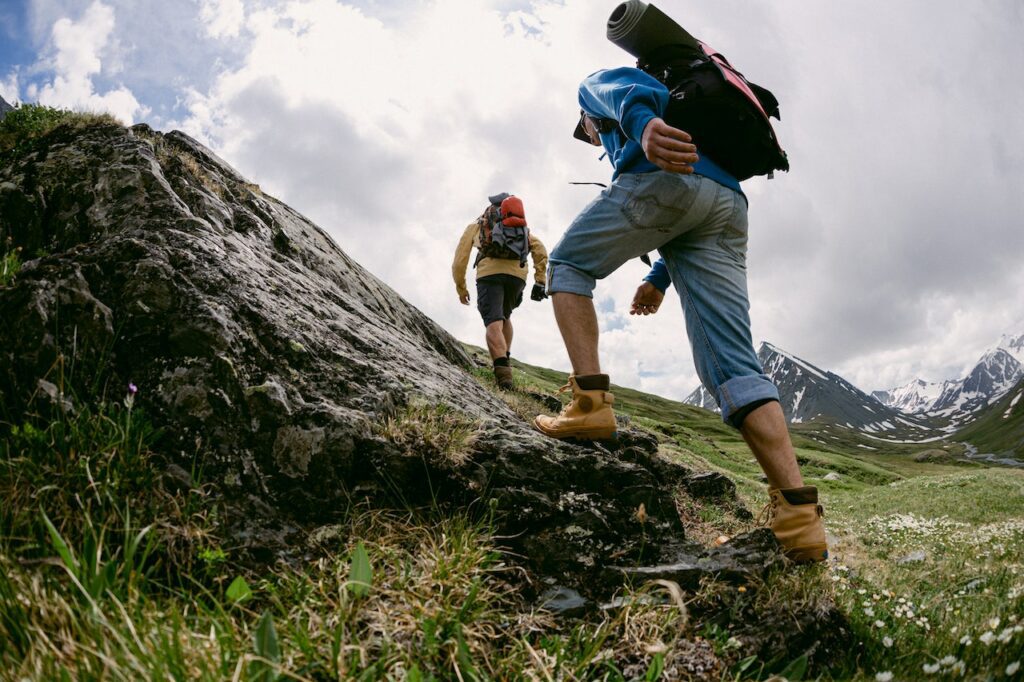 Two men climbing a green hill while wearing two pairs of hiking boots.