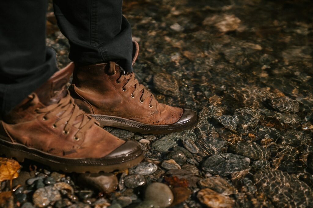 Man wearing black pants with a pair of brown hiking boots on shallow water with rocks.